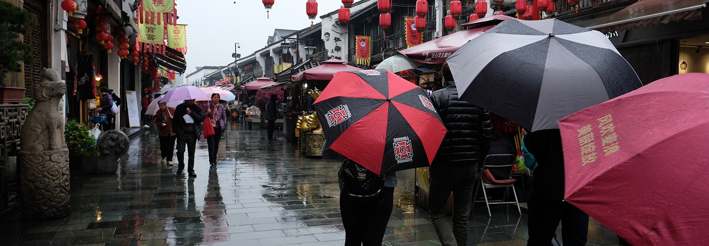 Rainy street scene with many people shielded by umbrellas. In the center of the scene is a person with a red and black umbrella with the honor G
