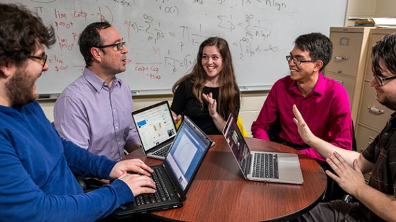older man talks to younger people, three laptops on a table between them
