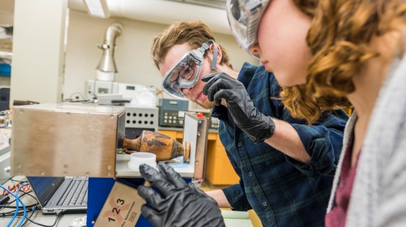 Nora and Ben place a greek vase in a spectrometer machine