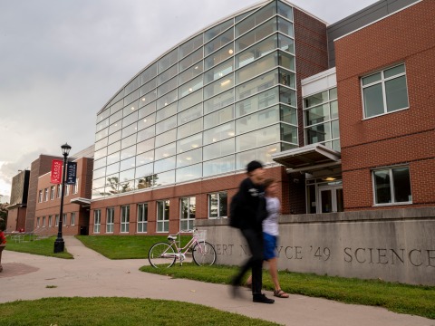 Two blurred figures walk in front of the entirely glass facade of the Noyce Science Center.