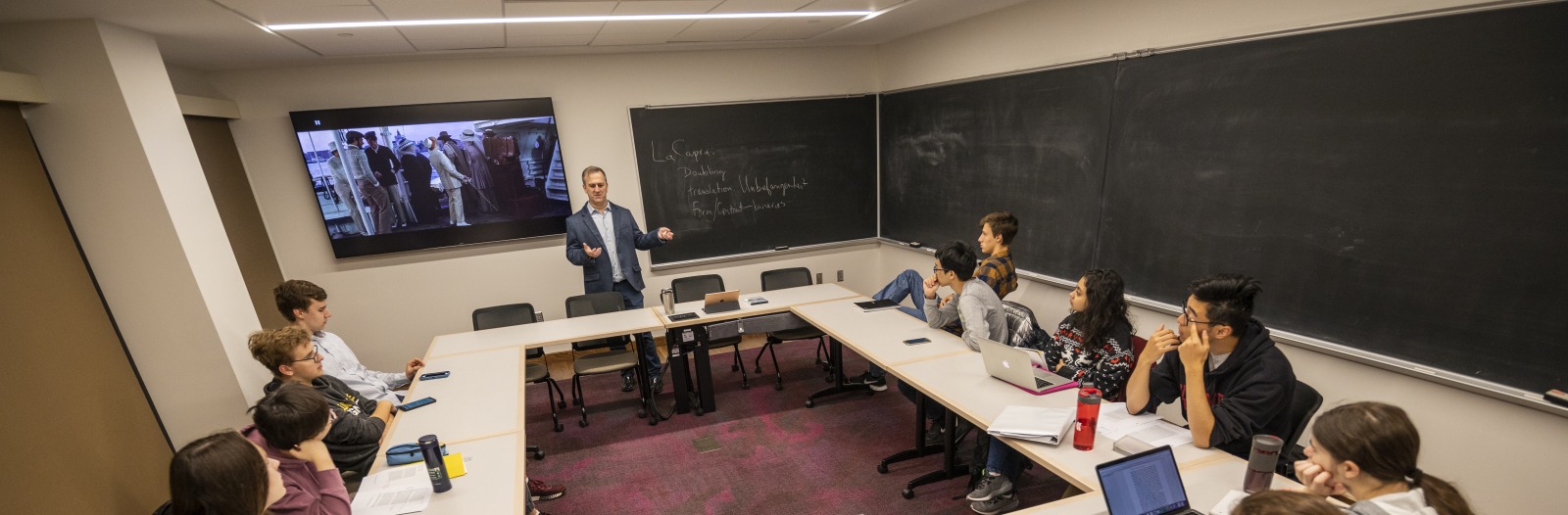 Man stands in front of a screen and blackboard with students in a horseshoe around him
