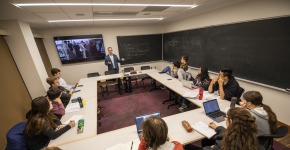 Man stands in front of a screen and blackboard with students in a horseshoe around him