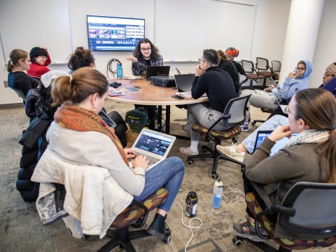 About a dozen students and instructor listen to another student presenting informally in their classroom