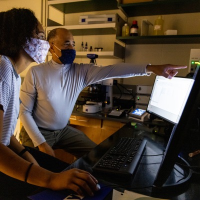 Pascal Lafontant points to a computer screen, while a student follows his gesture. Both wear face masks. They are in his laboratory.