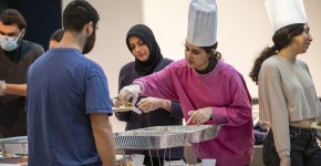 A young woman in a tall white chef's hat serves food to people waiting in line.