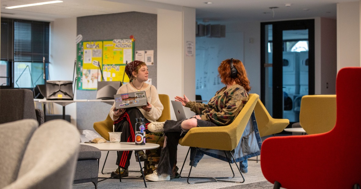 two people sit in large chairs in a lounge with a bulletin board and windows in the background