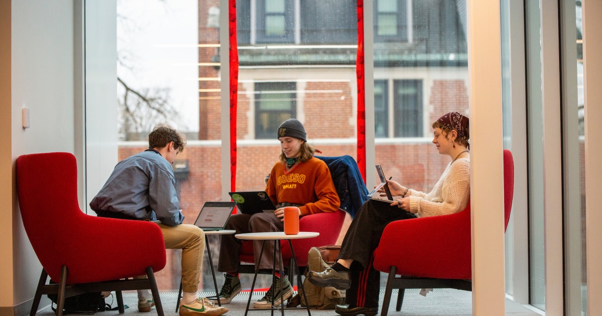 Three people sit in colorful chairs around a small table in a sunlight area with ceiling height windows