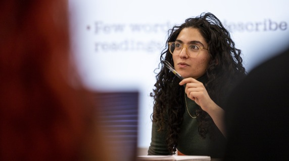 A woman in a green turtleneck and glasses looks intently off camera. In the foreground are the backs of students.