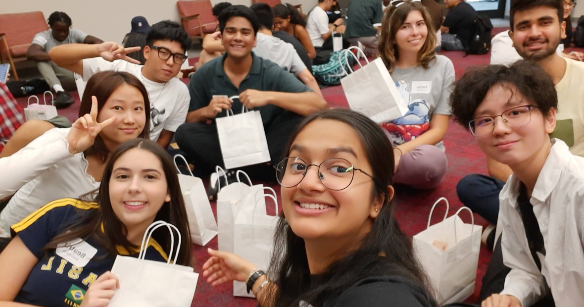 A small group of people sit on a red floor holding white bags and smiling at the viewer