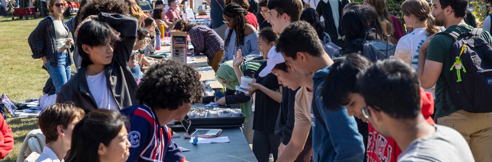 Crowded outdoor fair with a long line of students on either side of tables. Several are signing up for different organizations.