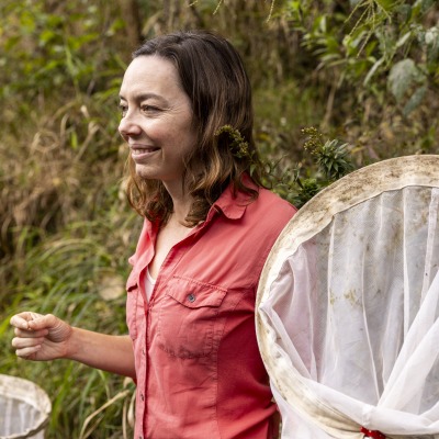 A woman wears a pink button down shirt and stands in a river with a large butterfly net.