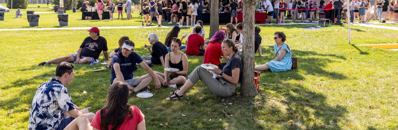 Family members, including student picnic under a shade tree on campus. Food tents and many people are visible in the background