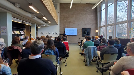 People are seated in front of a speaker, who wears a sweater and glasses. He is turned towards the presentation on the screen next to him.