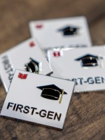 Four square white enamel pins sit on wooden planks The pins each have the word First-Gen and a mortar board in black with Grinnell College's red laurel leaves in upper left corner