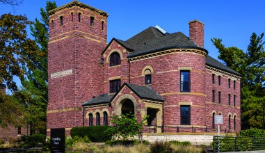 A full color photo of Goodnow Hall shows the reddish color of the rough-cut stone blocks and the highlights of tan stone. The roof of the square turrent is gone.