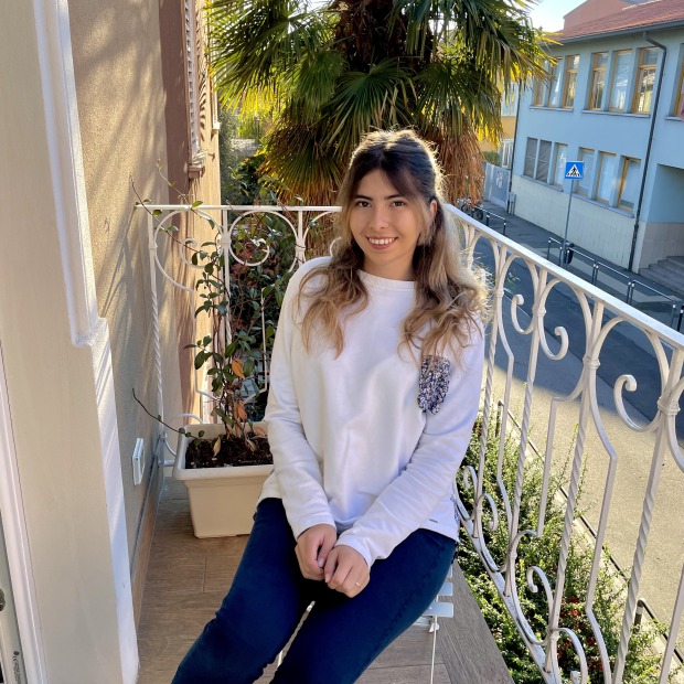 Emily sits in a chair on a balcony surrounded by plants and trees, smiling at the viewer