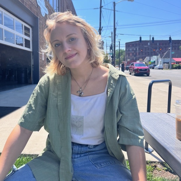 Gabriela sits outside in front of a street and tall brick building, smiling at the view