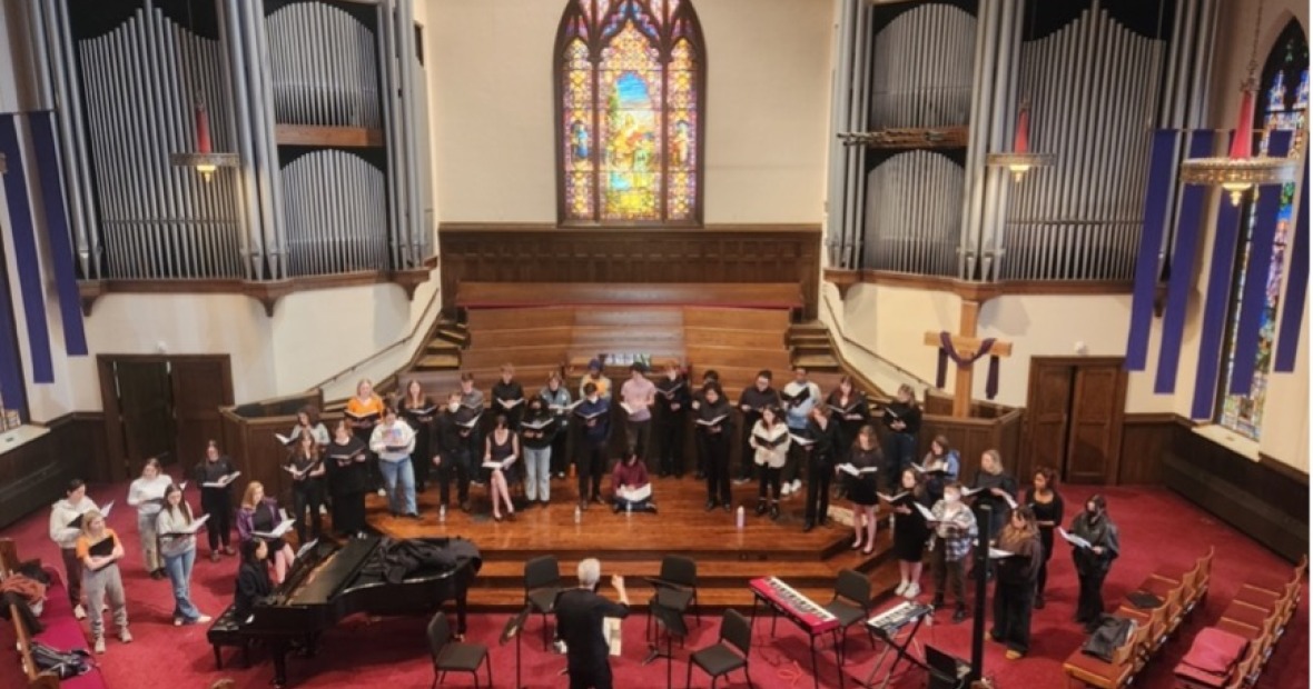 Singers in street clothes arrayed on a wooden dias in front of staine glass window and pipe organ