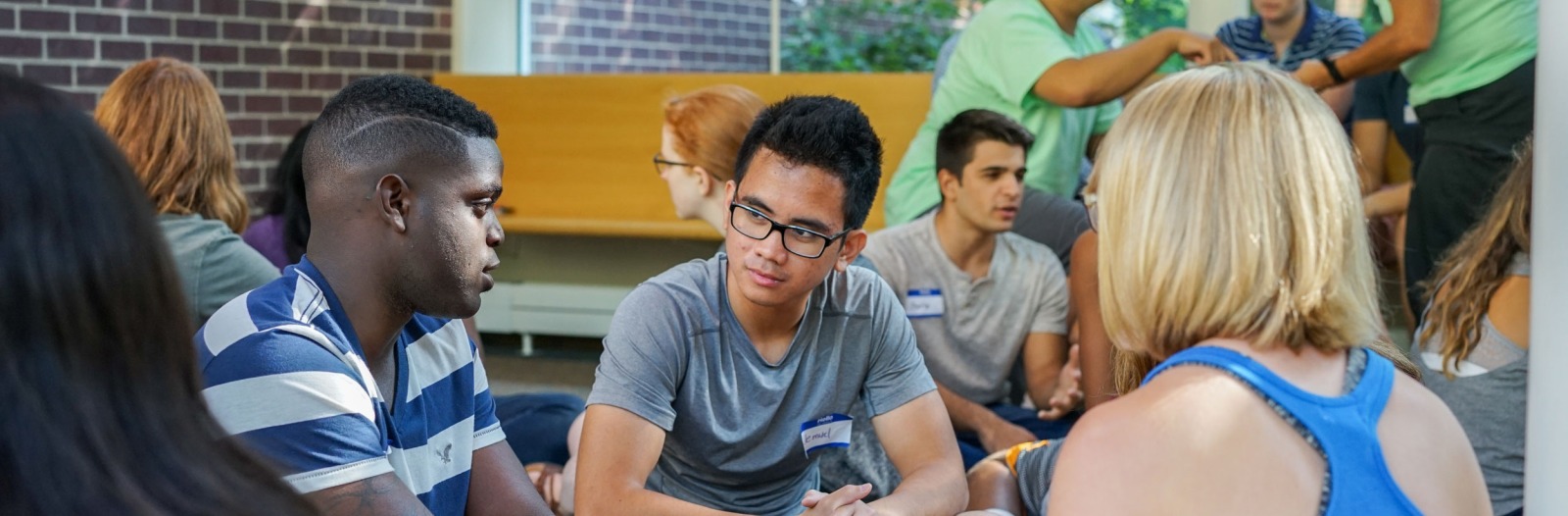 A variety of students gathered in a lounge. Several sit a a table, while others stand near a window