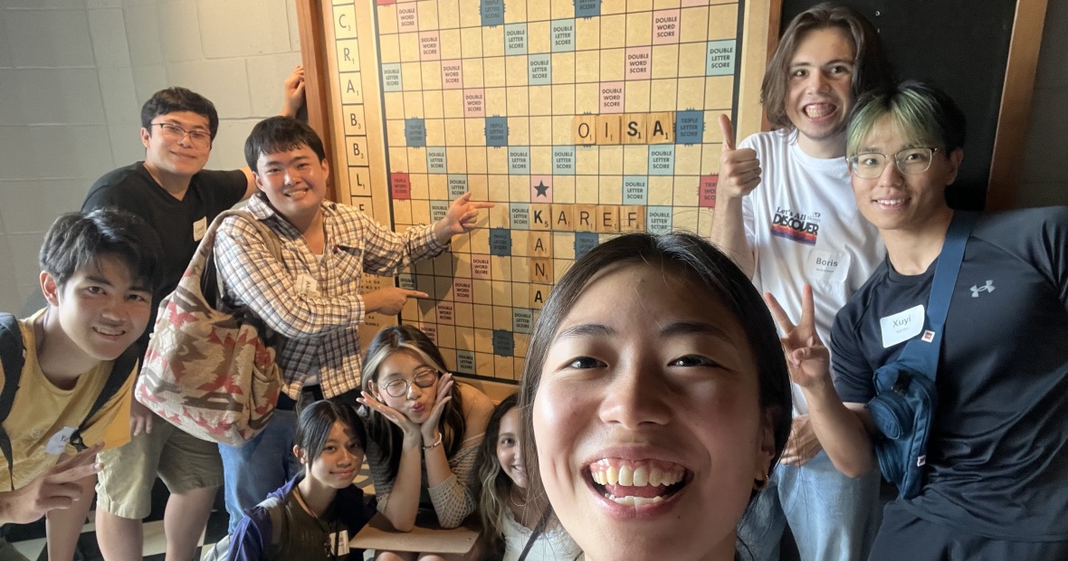 A group of people smile at the viewer while standing in front of a large Scrabble board where the letters "OISA" are written