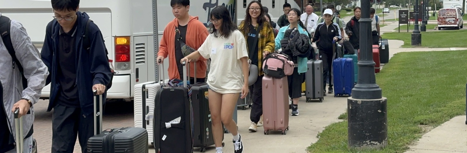 Many people walk towards the viewer, away from a large white bus, and carry luggage with them