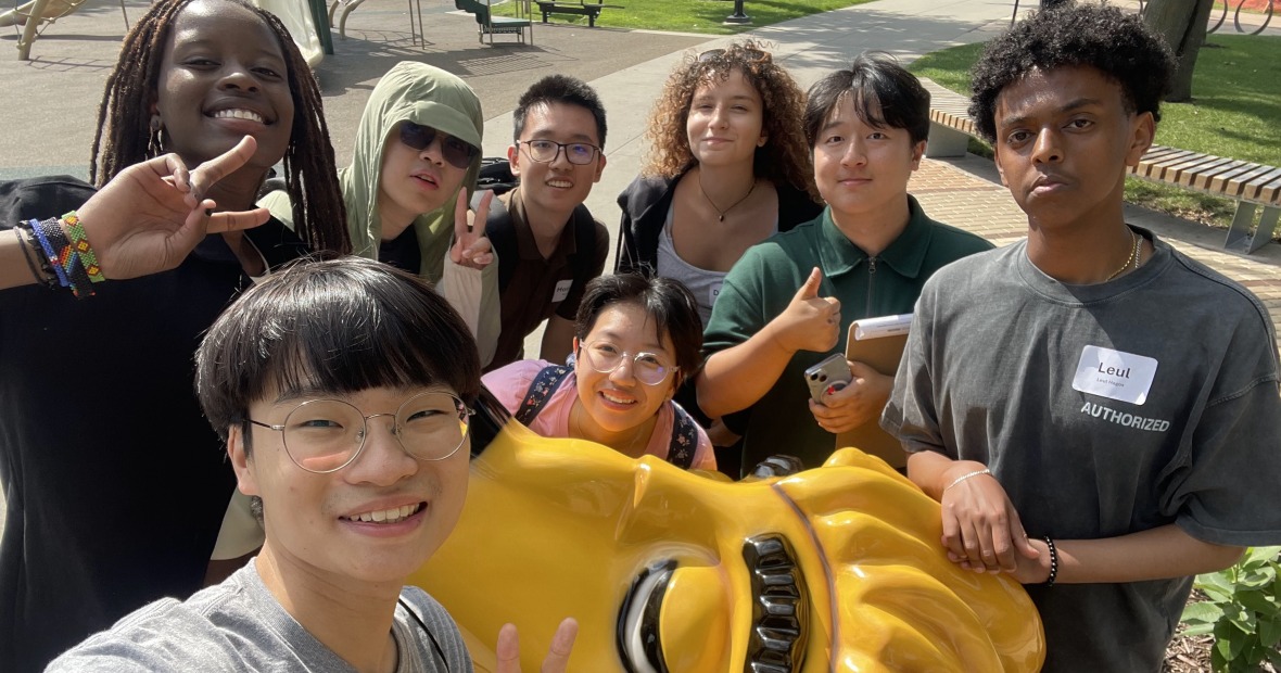 Several people stand in front of a yellow lion smiling at the viewer and holding up peace signs