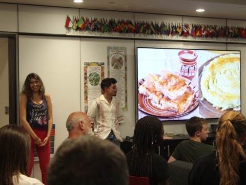 Two students smile as they present. A TV screen displays Bulgarian food.