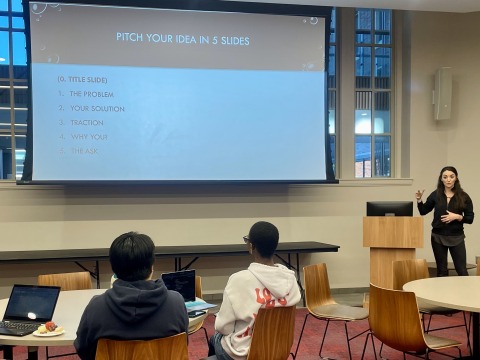 Andrea, at podium to the side of a slideshow, talks to tables of students. 