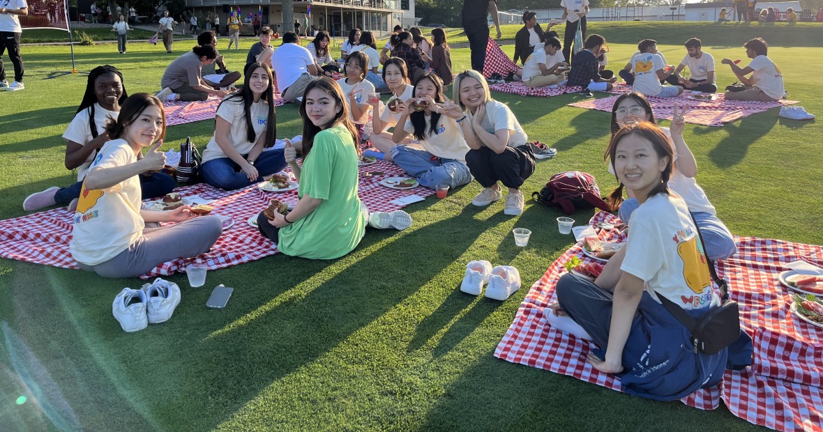 A group of people sit on red and white checkered blankets in the grass, hold plates of food, and smile at the viewer