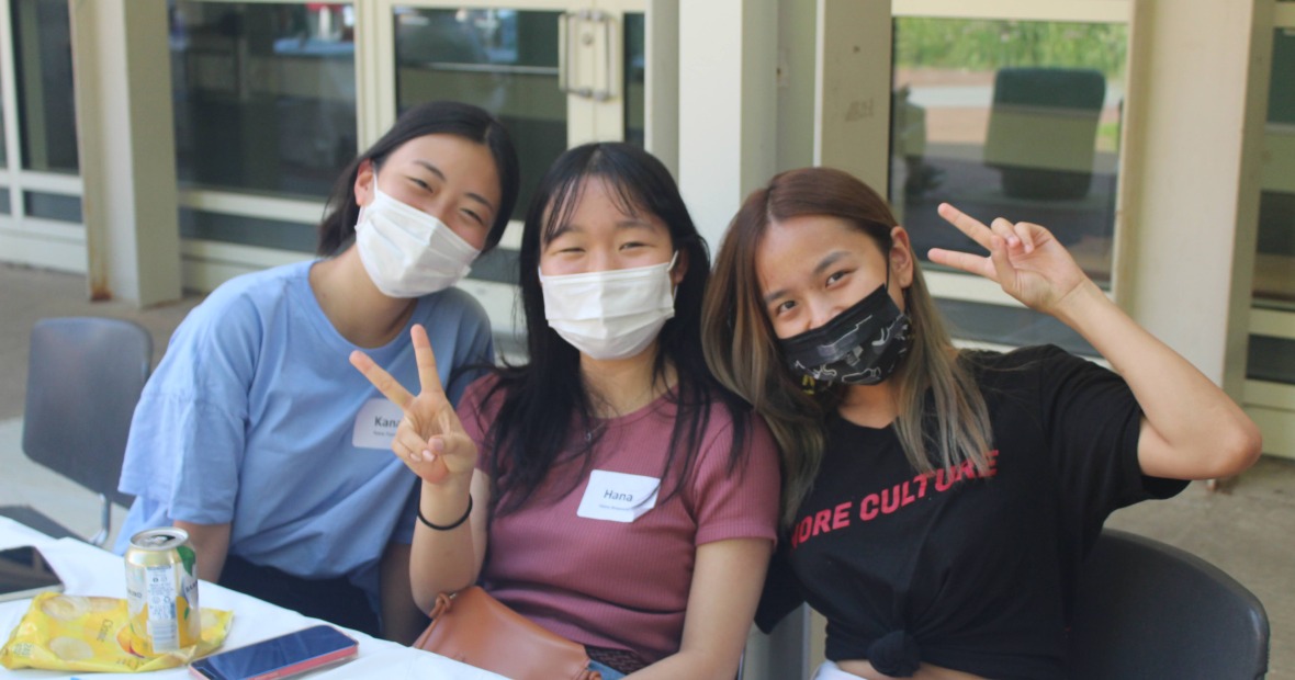 Three masked people sit at a table and flash the viewer with peace signs