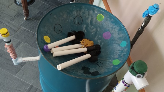 A large blue barrel, PVC pipe pieces, wood, and fabric are repurposed to make a musical instrument.