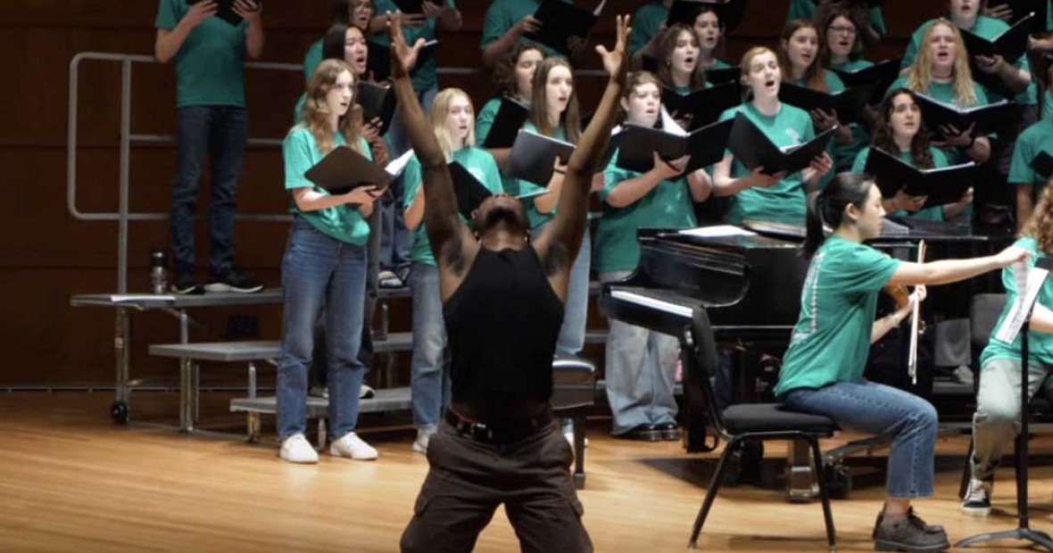 Black man in black tank, with arms raised and head thrown back, stand in front of a choral group in green tees and jeans