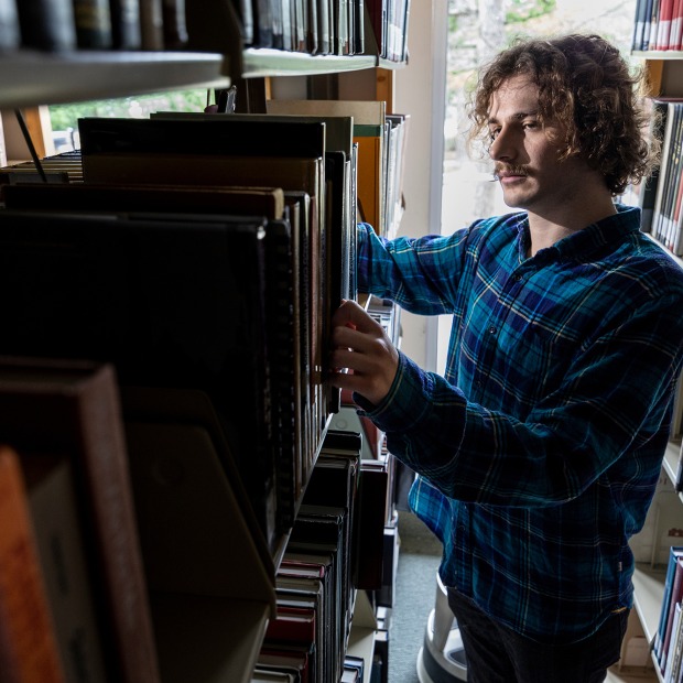 A young man stands in the library stacks examining a book
