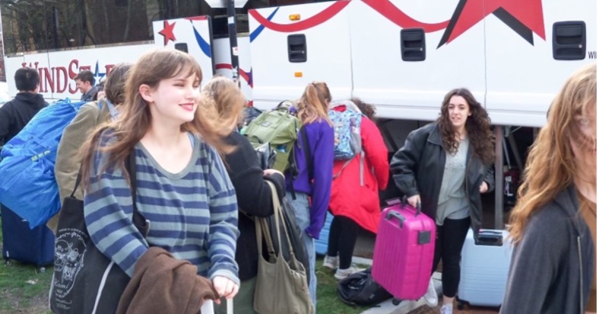 A large group of people in street clothes, many with luggage, stand around in front of a charter bus that has its storage panels open