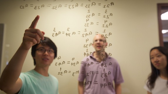 professor and students write equations on markerboard