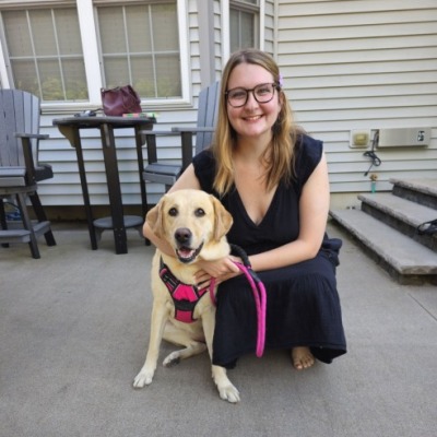 Mykenzie Masters, a blond woman with glasses, wears a dark dress. She is kneeling next to a yellow dog who is wearing a red and black harness with a pink leash