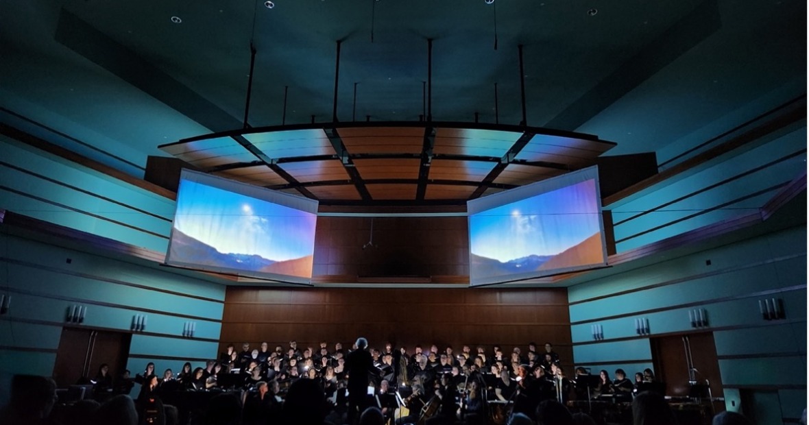 musicians in a dimly lit theatre, arrayed below two screens that display an outdoor image