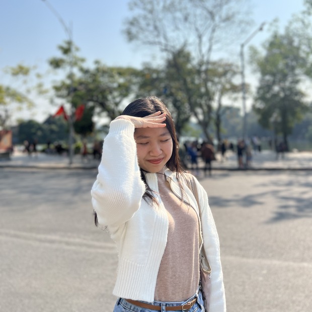 Phuong stands outside in front of a street lined with trees, smiling while looking down and her hand shading her face