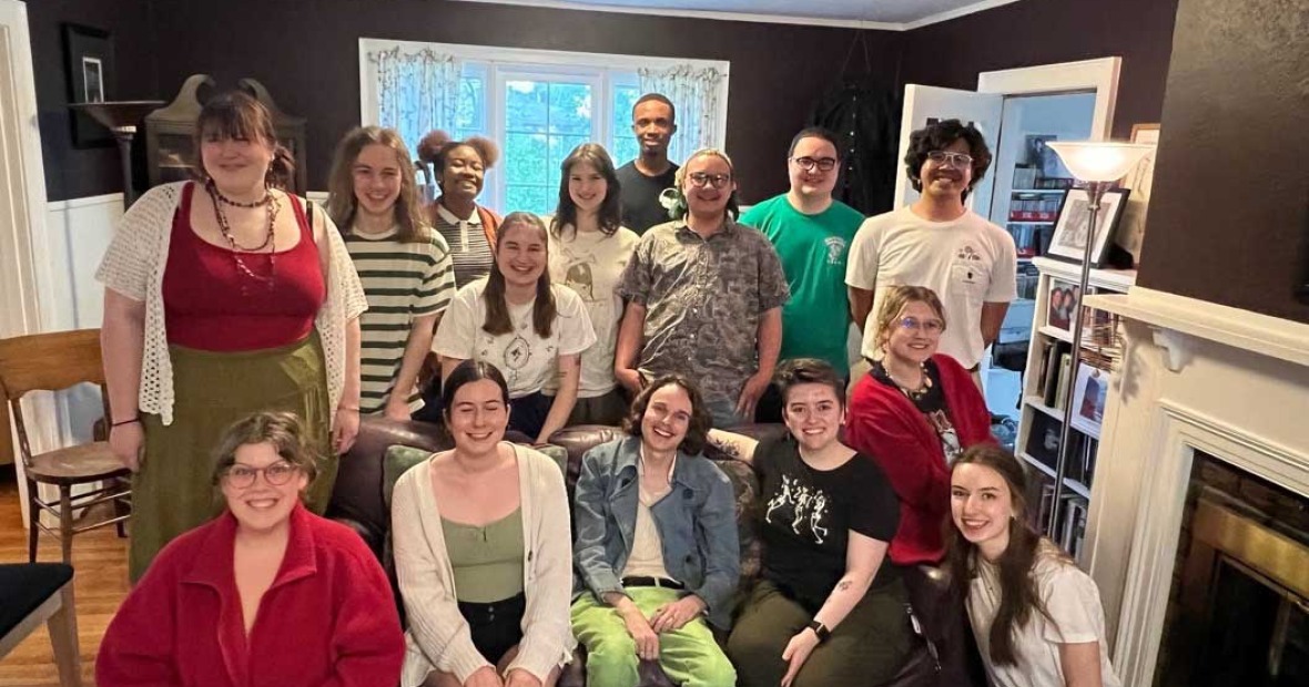 15 students, some seated on a couch, some crouched next to them, and others standing, in what appears to be a living room with a fireplace