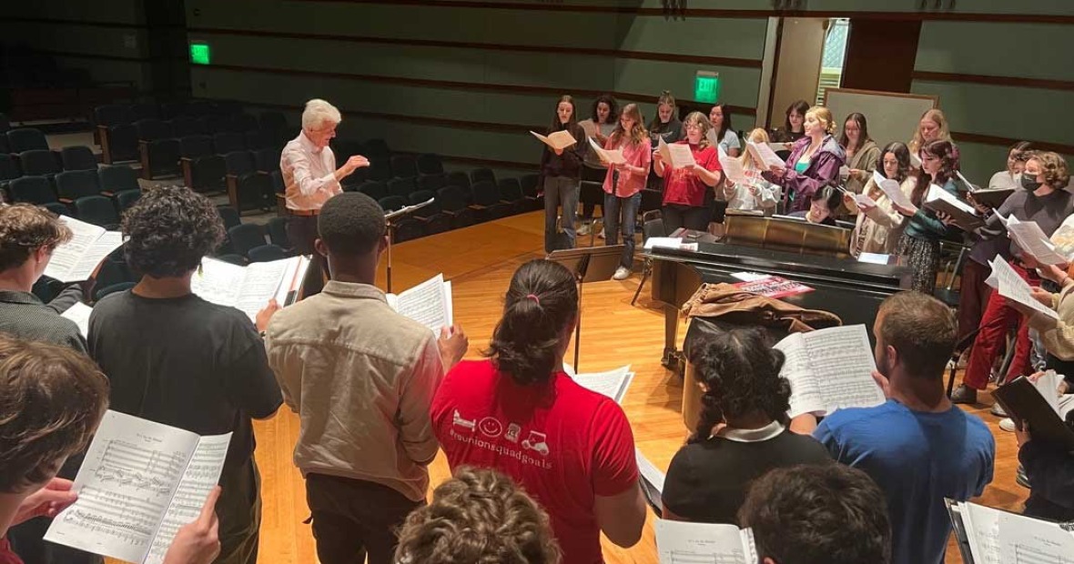 on a wooden stage, a man with white hair leads a choral group that stands arrayed in a horseshoe in front of him