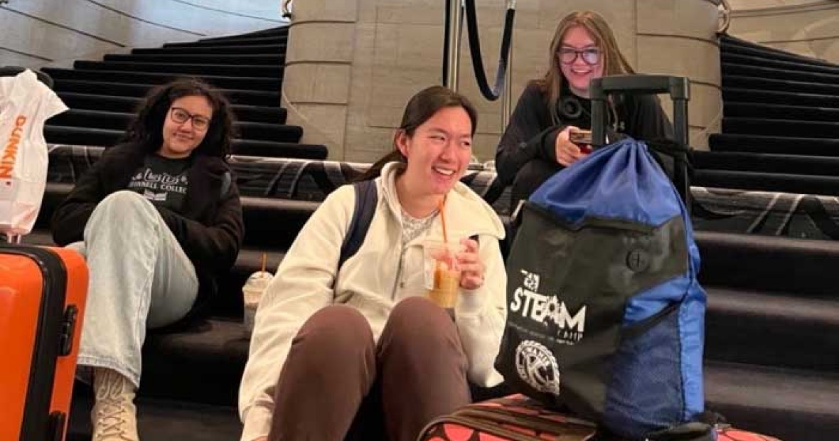 Three students sit on carpeted steps suitcases in front of them, an ornate lobby appears above and behind them.