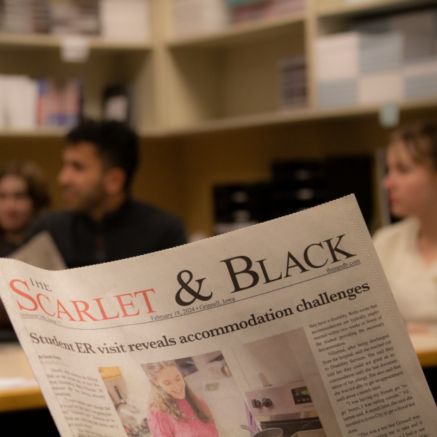 A closeup of the front page of the Scarlet and Black newspaper, while newspaper staff discuss the issue in the background.