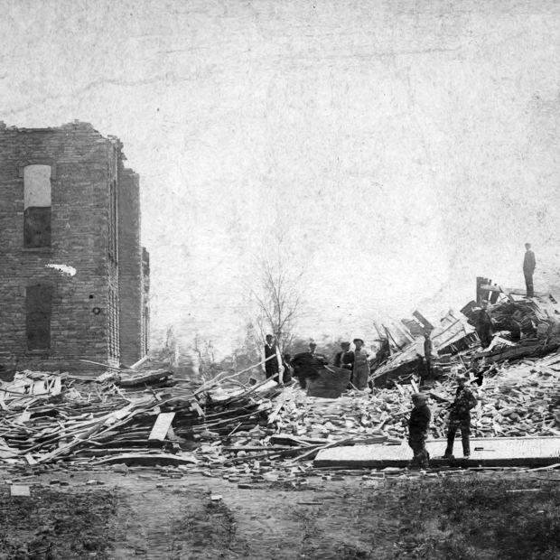 Several onlookers view two stories of a wrecked brick building that lies next to an enourmous pile of rubble