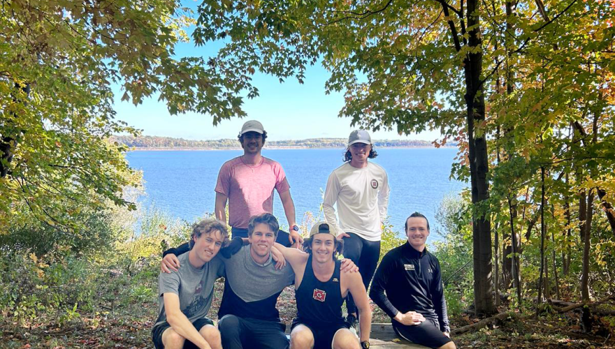 Brian and five friends pose on a picnic table with leafy trees and a lake in the background.
