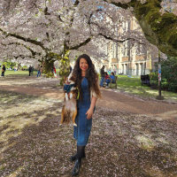 Cassey stands under a blooming cherry blossom tree and holds her dog. She is wearing a denim dress and has her long hair down.