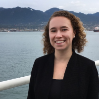 Caroline stands in front of the Pacific Ocean, with mountains in the background. She wears a black shirt and is smiling at the camera.