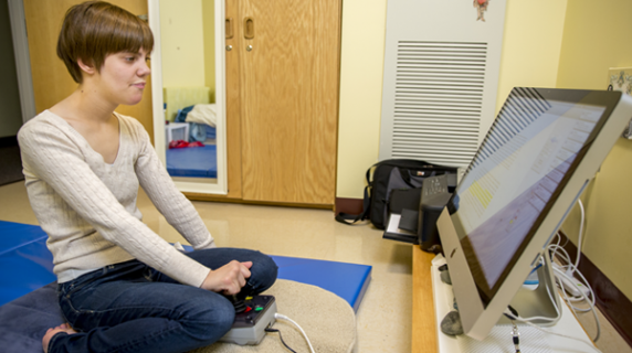 Jennifer Brooks ’15 sits on a blue gymnastics mat, facing a Mac on a table about 4 inches off the floor.