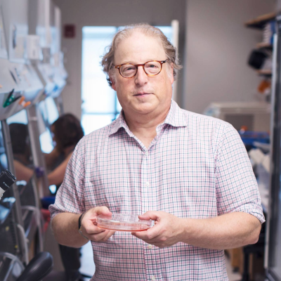 Dr. Landau stands in his lab and holds a petri dish. He is wearing glasses and a light purple button down shirt.