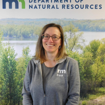 A woman wearing a Minnesota Department of Natural Resources sweater stands in front of a lake and smiles at the camera.