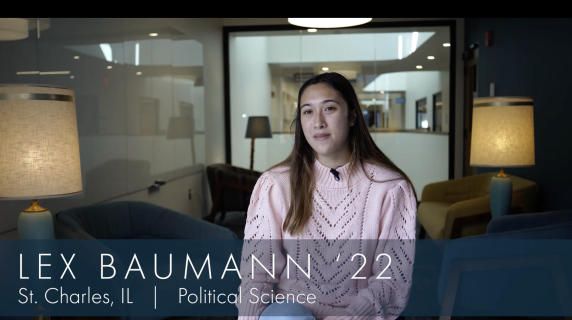 Lex Baumann, Political Science major from St.Charles, IL looks into the camera in a college study area. There are chairs, lamps, and a glass wall that reveals the rest of the building.